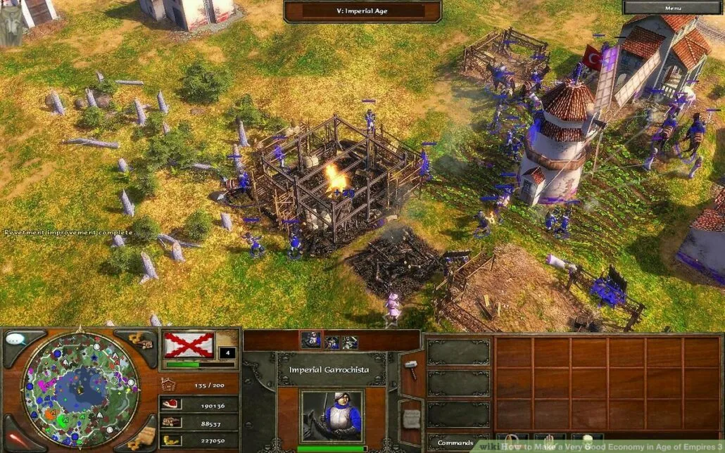 Captura Age Of Empires III Definitive Edition – Varios Aldeanos Construyen Un Edificio En Llamas Mientras Otros Cultivan Y Trabajan Cerca De Molinos