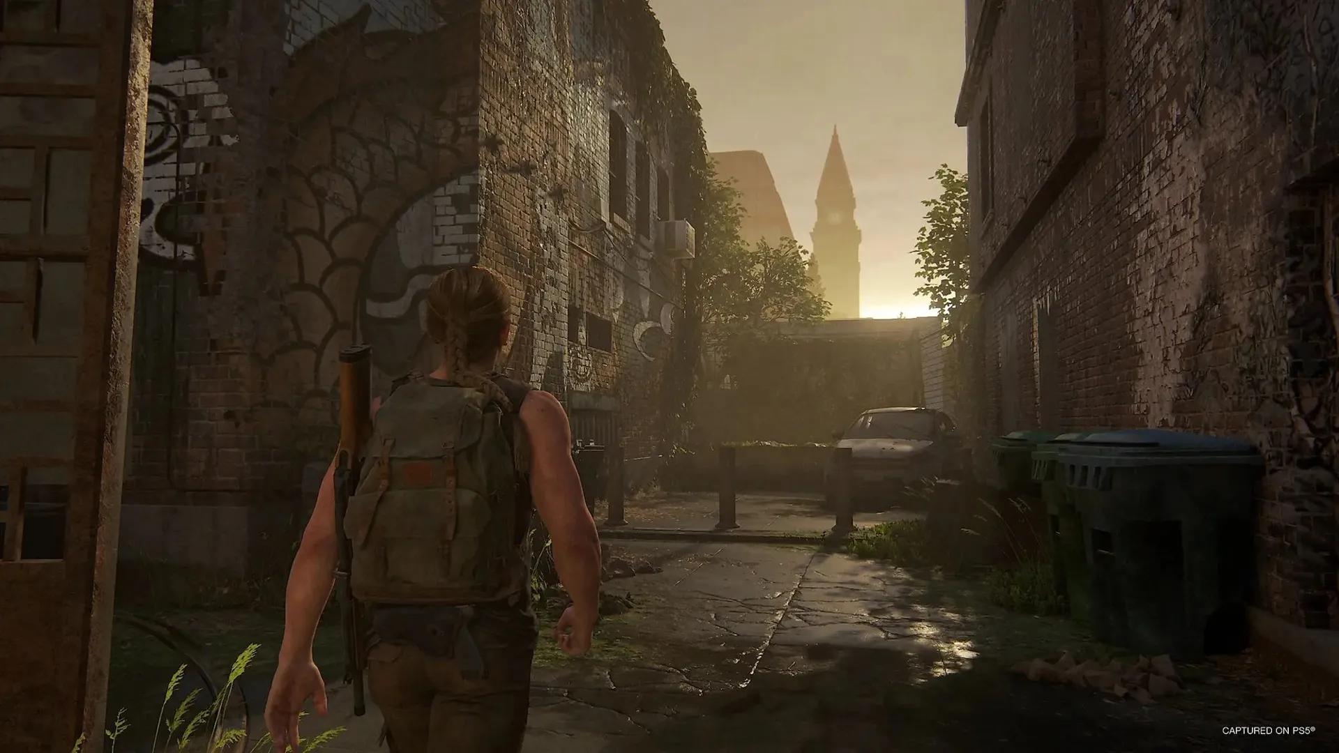Captura The Last Of Us Part II Remastered – Abby Camina Por Un Callejon Invadido De Vegetacion Entre Muros Grafiteados Con Un Coche Abandonado Al Fondo